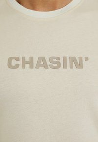 Kerman puuvillapaita, jossa on kohokuvioitu vaaleanbeige teksti "CHASIN'" edessä. Kaula-aukko on hieman kontrastivärinen vaalea.