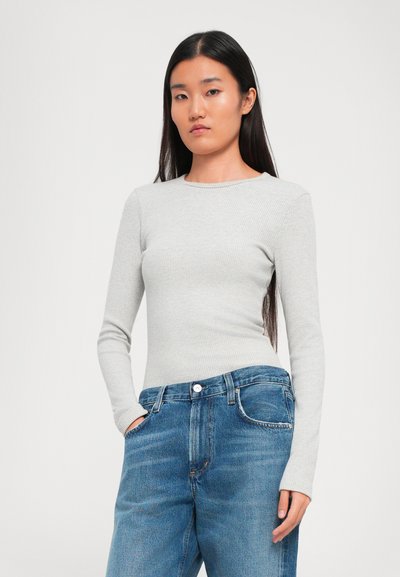 Woman with long dark hair wearing a light grey ribbed long-sleeve top and blue denim jeans, standing with one hand in pocket.
