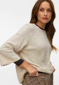 Femme aux longs cheveux bruns portant un pull beige par-dessus un haut foncé et une jupe à carreaux, ajustant la jupe de la main.