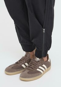 Smeđe Adidas tenisice s bijelim prugama nosene uz crne hlače s patentnim zatvaračem na gležnju.