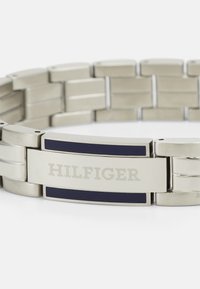 Rozsdamentes acél karkötő váltakozó fényes és matt láncokkal, középen található csattal, amelyen gravírozott "HILFIGER" felirat és tengerészkék díszítés van.
