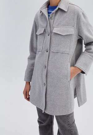 Person trägt einen hellgrauen, geknöpften Wollmantel mit Klappentaschen auf der Brust über einem blau gestreiften Pullover und dunkelgrauen Jeans.