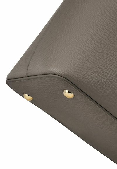 Coin inférieur d'un sac à main en cuir taupe texturé avec deux pieds en métal doré visibles.