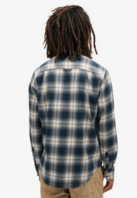 Camisa de franela de cuadro en azul, crema y marino, con mangas largas, dobladillo curvado y puños abotonados. La parte trasera tiene un cuello estándar y un detalle de logo.