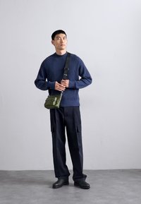 Pulover ribat cu guler în navy, pantaloni cargo negri lejeri și pantofi negri. Geantă mică de umăr în verde măsliniu cu detalii cu fermoar. Design minimalist.