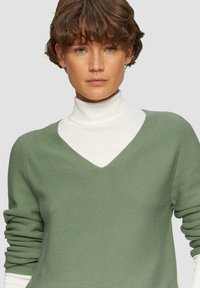 Groen gestructureerd sweater met een V-halsontwerp, gelaagd over een witte coltrui. De stof lijkt zacht en comfortabel.