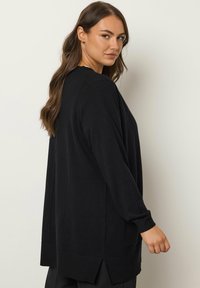 Schwarzer Langarm-Cardigan mit lockerer Passform, seitlichen Schlitzen am Saum und gerippten Bündchen. Das Material wirkt weich und hat eine glatte Textur.