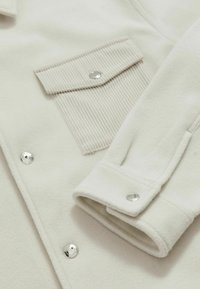 Chaqueta de forro polar blanca con un bolsillo en el pecho de canalé, botones de presión plateados y un cuello estructurado. Textura suave con un diseño limpio y minimalista.