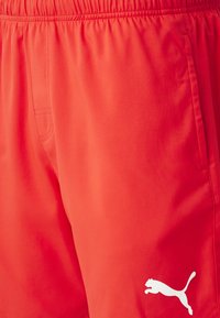 Sarkanas sporta šorti, izgatavoti no gludas auduma, ar elastīgu jostasvīru un baltu Puma logo zem kreisā sāna.