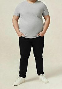 Persoon staat met handen in de zakken, draagt een grijs shirt met korte mouwen, zwarte broek en witte sneakers tegen een effen achtergrond.