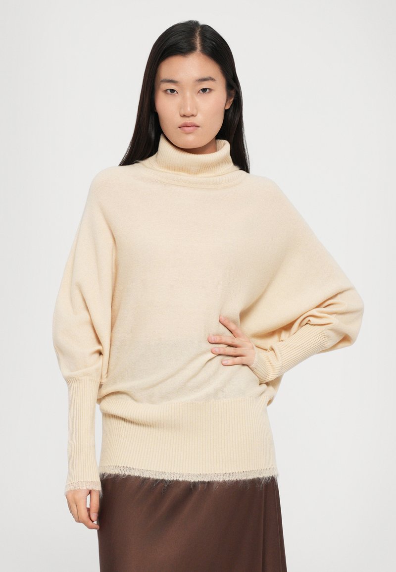 Pull encolure roulée crème avec une texture tricotée lâche, des poignets côtes et un ourlet effiloché. Stylé sur une jupe marron, avec une coupe décontractée.