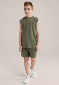 Ärmlös olivgrön bomullstopp med axeldetalj och broderad logotyp, i kombination med matchande shorts och vita sneakers.