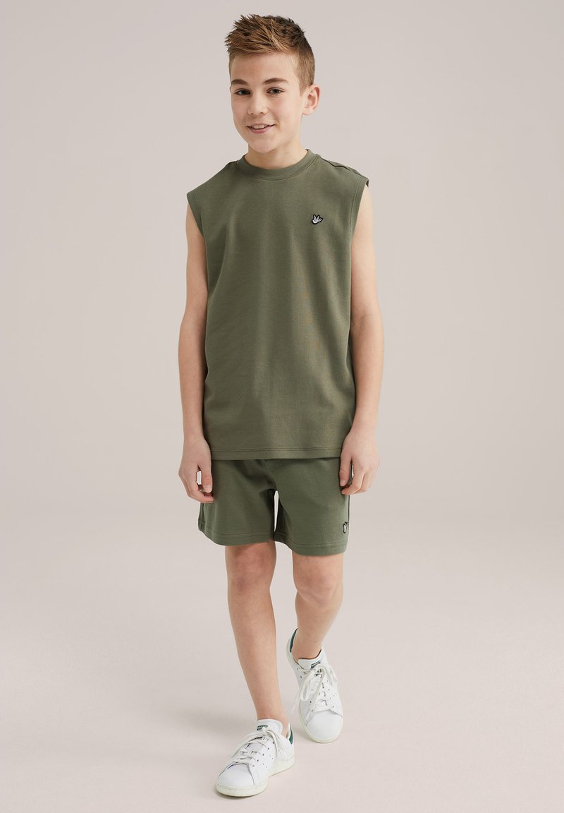 Ärmlös olivgrön bomullstopp med axeldetalj och broderad logotyp, i kombination med matchande shorts och vita sneakers.