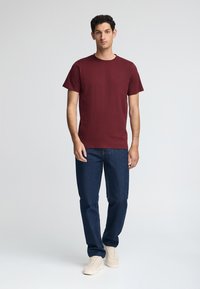 Bordeaux kortegalm T-shirt met een ronde hals en een klein logo, gecombineerd met donkerblauwe spijkerbroek en beige sneakers. Soepele textuur, casual pasvorm.