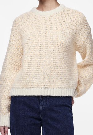 Person trägt einen cremefarbenen, strukturierten Strickpullover mit gerippten Bündchen und Saum, kombiniert mit dunkelblauen Jeans.