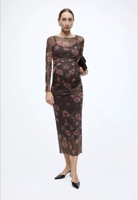 Langermet, tettsittende kjole i gjennomsiktig brunt stoff med rosa blomstermønster, rynkedetaljer og midilengde. Brukt med svarte tilbehør.