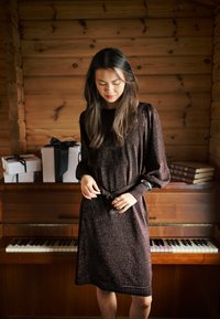 Robe marron à manches longues scintillante avec une taille ceinturée, présentant un col rond et un tissu texturé. En arrière-plan, un piano et des boîtes-cadeaux.