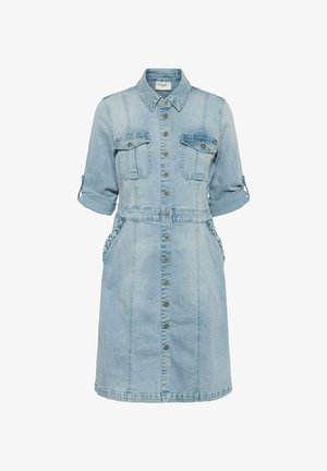 Világoskék farmer inget visel, gombos lezárással, két mellzsebbel, felhajtott ujjakkal és oldalán texturált díszítésű zsebekkel.