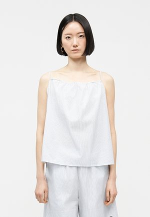 Νέα γυναίκα με κοντά μαύρα μαλλιά, που φοράει λευκό αμάνικο τοπ με λεπτές ρίγες και χαλαρό παντελόνι που ταιριάζει, κοιτάζοντας μπροστά σε απλό φόντο.