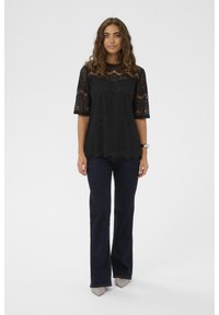 Fekete csipke felső rövid ujjakkal, bonyolult mintákkal, réteges kialakítással és ívelt szegéllyel, sötétkék, trapéz farmernadrággal párosítva.