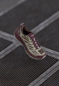 Scarpa da corsa leggera con tomaia beige, accenti bordeaux, punta testurizzata e dettagli riflettenti; presenta una suola spessa con trazione.