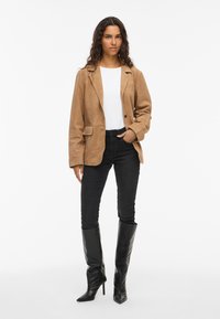 Beige suede blazer with lapels, single button closure, and side pockets, paired with black skinny jeans and knee-high black stiletto boots.