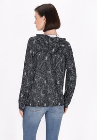 Jachetă cu glugă de culoare neagră cu modele florale albe, dintr-un material ușor, cu mâneci lungi și o croială lejeră, asociată cu blugi albaștri.