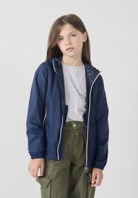 Jongere persoon die een marineblauwe hoodie over een lichtgrijze shirt en olijfgroene cargo-broeken draagt, staande tegen een effen lichte achtergrond.