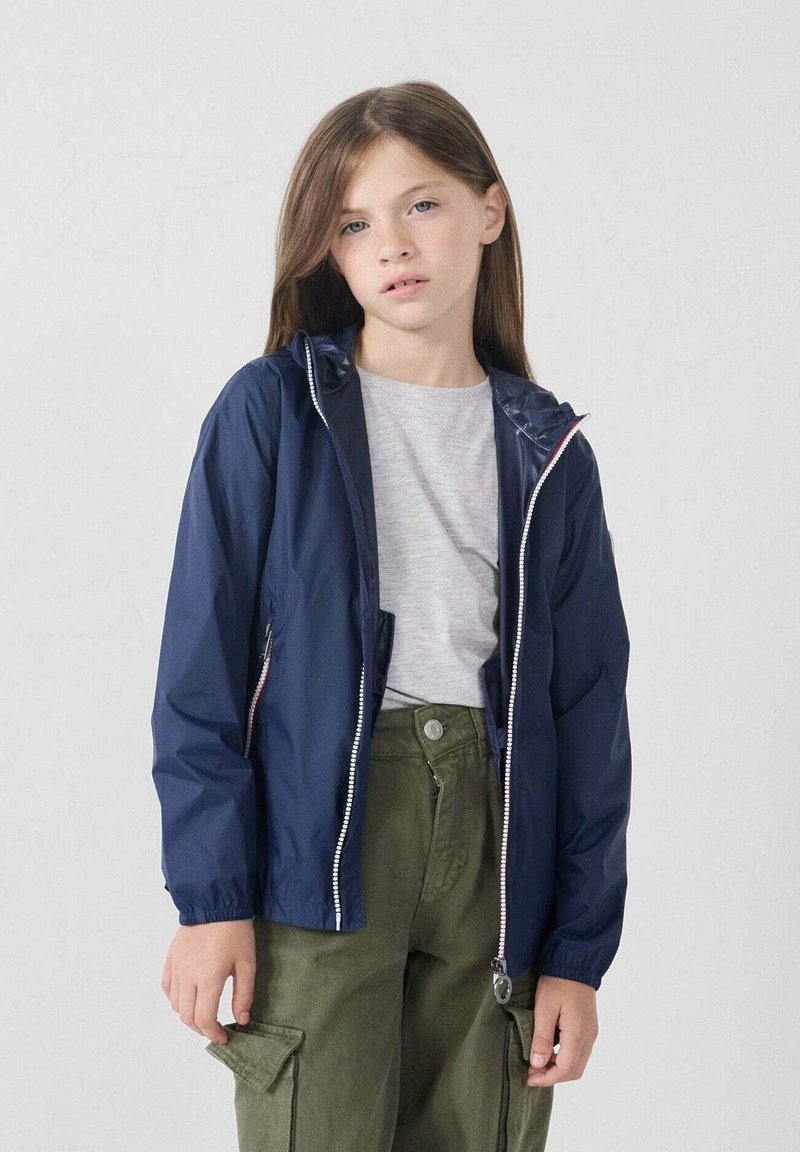 Jongere persoon die een marineblauwe hoodie over een lichtgrijze shirt en olijfgroene cargo-broeken draagt, staande tegen een effen lichte achtergrond.