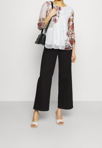 Blusa blanca de flores con mangas transparentes, combinada con pantalones de pierna ancha negros y sandalias de tacón ligero de color claro. Bolso cruzado negro.