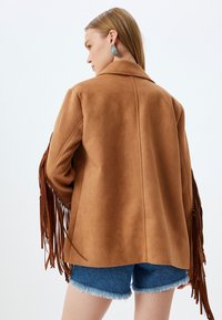 Bruine suède jas met franjes op de mouwen, getailleerde pasvorm en een gelapte kraag, gedragen met spijkerbroek shorts.