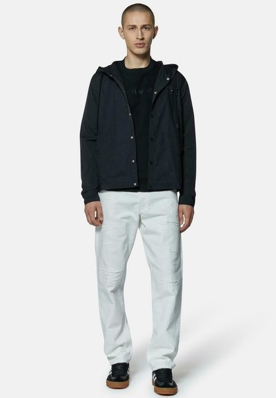 Giacca leggera nera con cappuccio sopra una maglietta nera, abbinata a pantaloni bianchi strappati e sneakers nere con suole marroni.