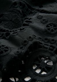 Schwarzer Stoff mit aufwändigen Ausschnittmustern und strukturiertem Stickereien, darunter florale Designs und kreisförmige Motive, die Tiefe und Detail schaffen.