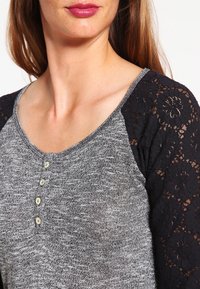 Top grigio mélange con maniche raglan in pizzo nero, scollatura arrotondata e chiusura con bottoni sul davanti. Tessuto morbido in tutto.