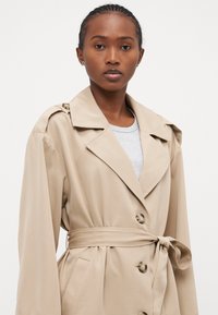 Beige trench coat med dobbelspent design, bred krage, belted midje og store knapper. Glatt stoff med sidelommer.