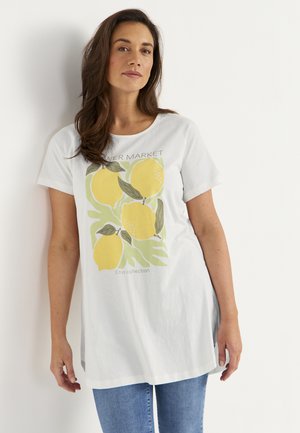 Frau trägt ein weißes übergroßes T-Shirt mit gelbem Zitronenmotiv und dem Schriftzug „Citrus Collection“, kombiniert mit blauen Jeans, vor einem einfarbigen Hintergrund.