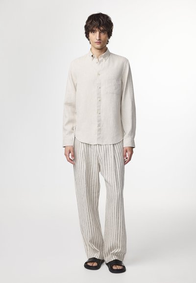 Lys beige linned skjorte med knapper og en enkelt brystlomme, parret med brede stribede bukser i lys beige og sort. Sorte sandaler.