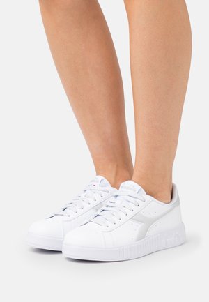 Zapatillas blancas bajas con cordones, usadas en piernas sobre un fondo liso y claro.