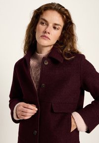 Woman with wavy brown hair buttoning a dark burgundy coat over a pink knit sweater, looking down and to her left against a plain background.