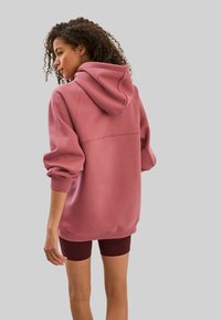 Sweat-shirt à capuche rose avec une coupe décontractée, des poignets côtelés et une texture lisse, associé à un short bordeaux foncé. Vue de dos.