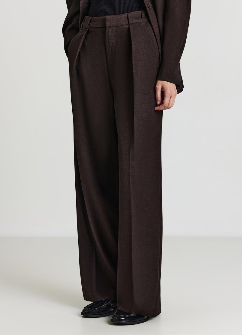 Pantalon à jambes larges marron en tissu lisse, avec une taille haute plissée. Associé à des chaussures noires à enfiler.