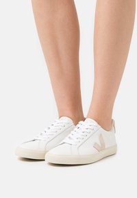 Sneaker bianche in pelle con lacci piatti, dotate di un logo in suede beige sul lato e di una suola in gomma texturizzata.