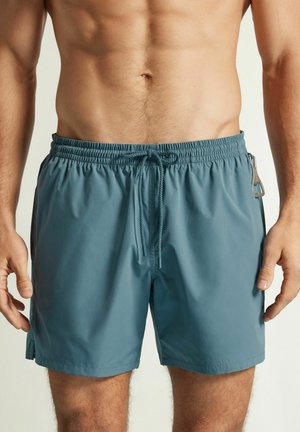 Męski tors i górna część nóg ubrani w niebieskie kąpielówki z elastycznym pasem, sznurkiem i małym breloczkiem przymocowanym po prawej stronie.