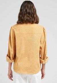 Chemise jaune moutarde en tissu texturé, avec des manches bouffantes, des poignets à revers et un dos droit.