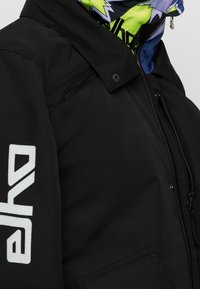 Veste noire avec texte blanc "elko" sur la manche, poches à fermeture éclair sur la poitrine et les côtés, et une capuche colorée verte et bleue en dessous.