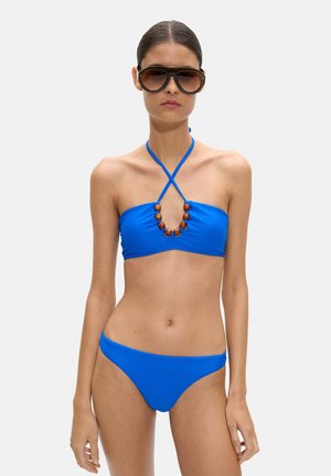 Kobieta w jasnoniebieskim bikini z drewnianymi koralikami na pasku przy szyi i dużych ciemnych okularach przeciwsłonecznych, stojąca na jednolitym tle.