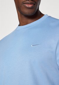Šviesiai mėlynas medvilninis marškinėliai su apvaliu kaklu ir trumpomis rankovėmis. Ant kairiojo krūtinės yra mažas sidabrinis Nike logotipas. Sklandus paviršius.