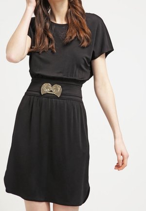 Femme portant une robe noire courte avec une large ceinture ornée d'une boucle métallique en forme de deux visages.