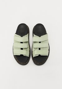 Zaļi zamšādas slaidu sandales ar trim Velcro siksnām, melnu gumijas zoli un kontrastējošu dzeltenu šuvumu malās.