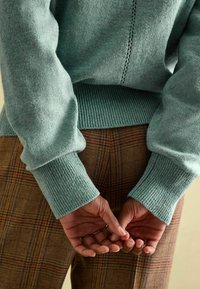 Pull en bleu sarcelle clair avec poignets côtelés, porté sur un pantalon marron à carreaux. Il présente une finition texturée et un détail de couture le long du dos.